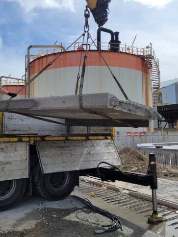 Una grúa está elevando una gran losa de hormigón sobre un camión en una zona industrial.