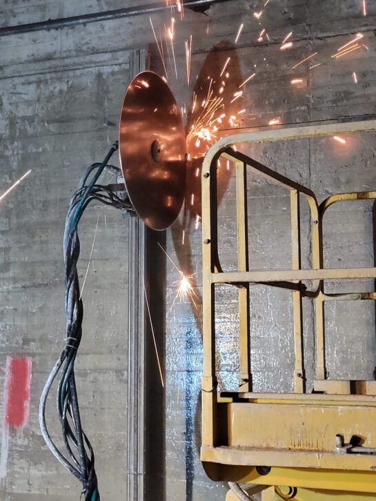 Un trabajador suelda una placa de metal desde una plataforma elevadora, con chispas que saltan contra una pared de hormigón.