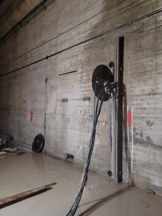 Muro de hormigón con un carrete de manguera montado y una manguera negra en un espacio industrial húmedo.