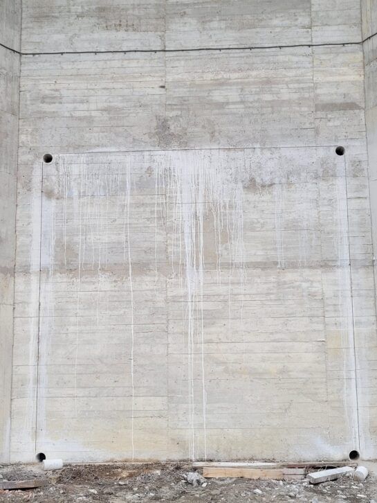 Muro de hormigón con un gran panel de color claro y pequeños orificios de montaje negros en las esquinas.