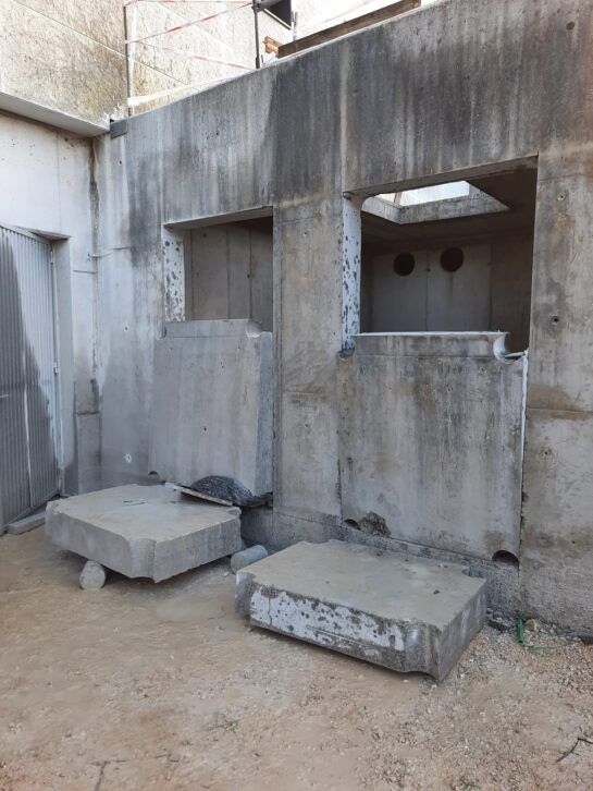 Muro de hormigón desgastado con dos pequeñas aberturas rectangulares y losas de hormigón sueltas en un patio polvoriento.