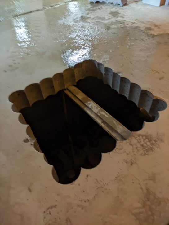 Escotilla abierta en el suelo con un tablón de madera sobre un suelo de hormigón mojado.