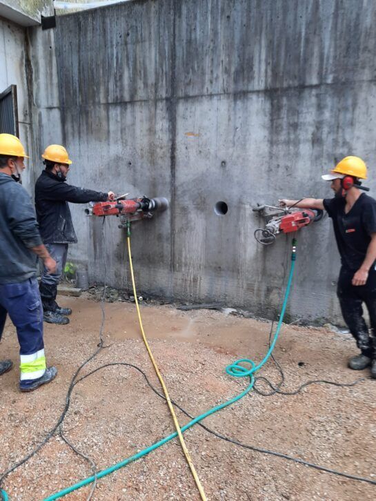 Trabajadores con cascos amarillos perforan una pared de hormigón con herramientas eléctricas con cable.