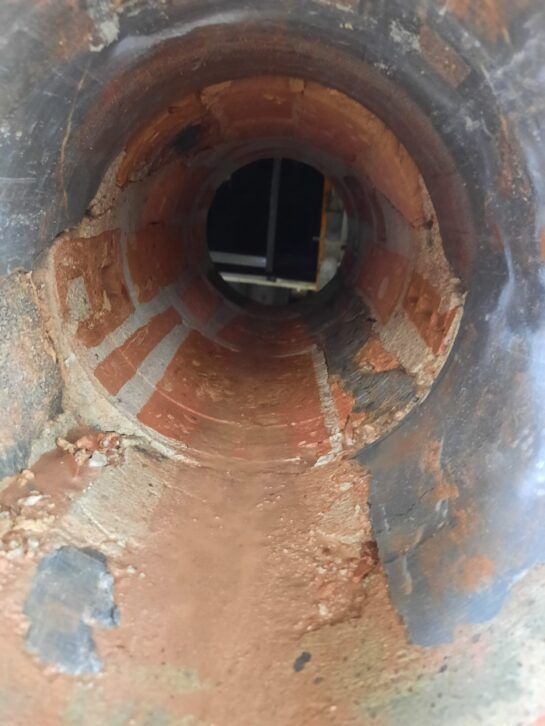 Interior de tubería metálica oxidada con paredes corroídas y escombros, vista desde el interior hacia el exterior.