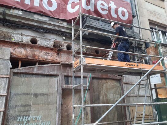 Fachada de un edificio en reparación, cubierta de andamios, con trabajadores y una pancarta roja que dice 