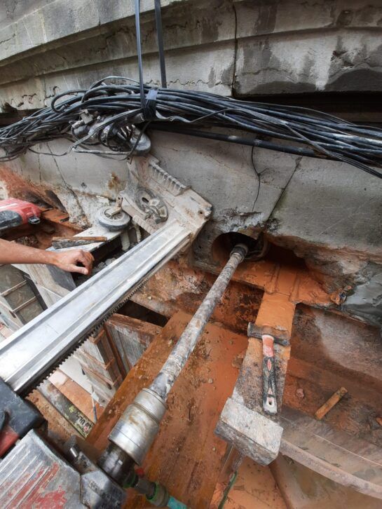 Un trabajador manipula cables en una zanja de hormigón dañada, con tuberías y vigas metálicas al descubierto.