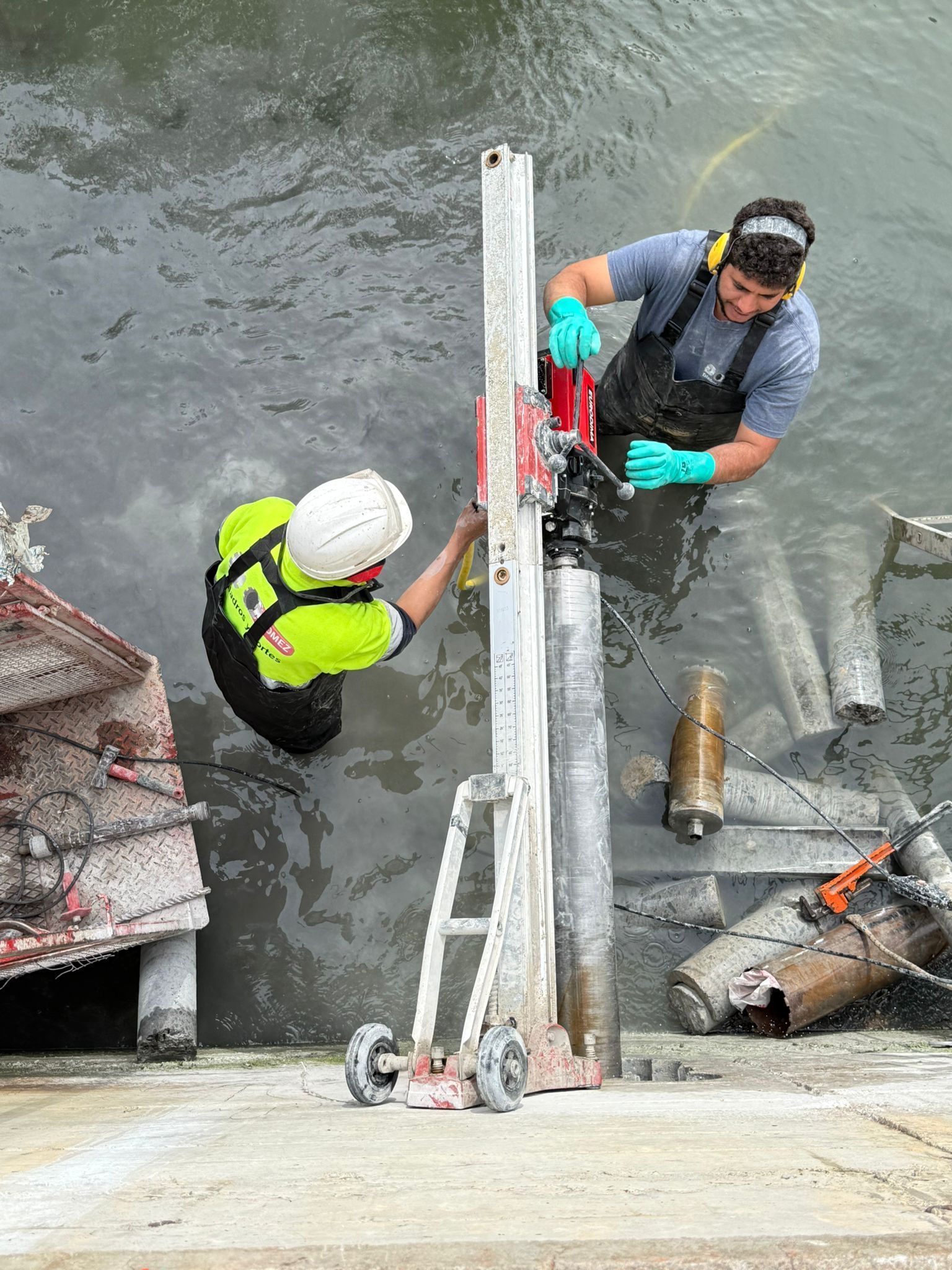 Dos trabajadores con equipo de seguridad manipulan cuerdas al borde de un muro de hormigón sobre el agua.