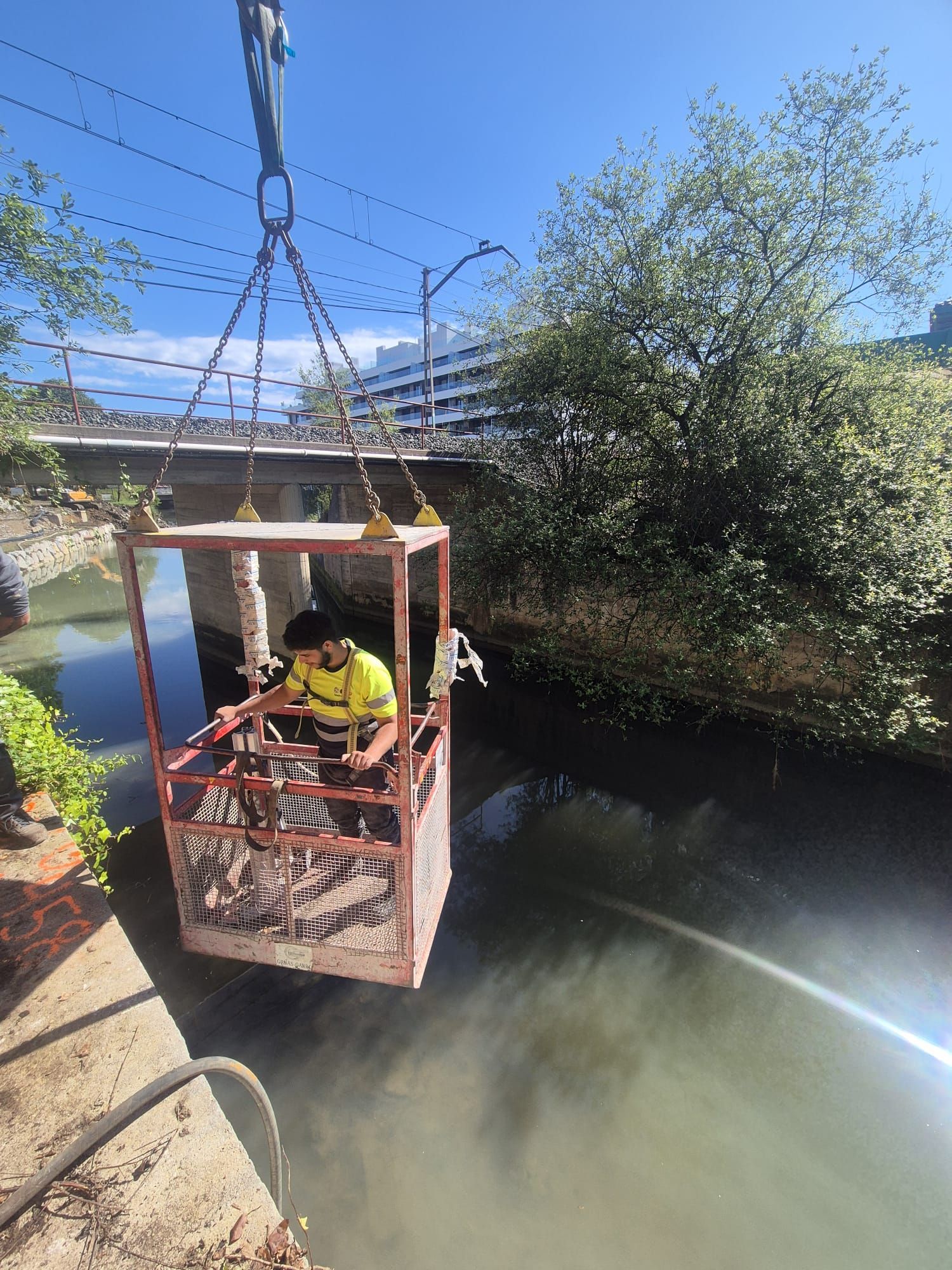 Trabajador con chaleco amarillo operando una plataforma suspendida sobre un canal cerca de un puente.