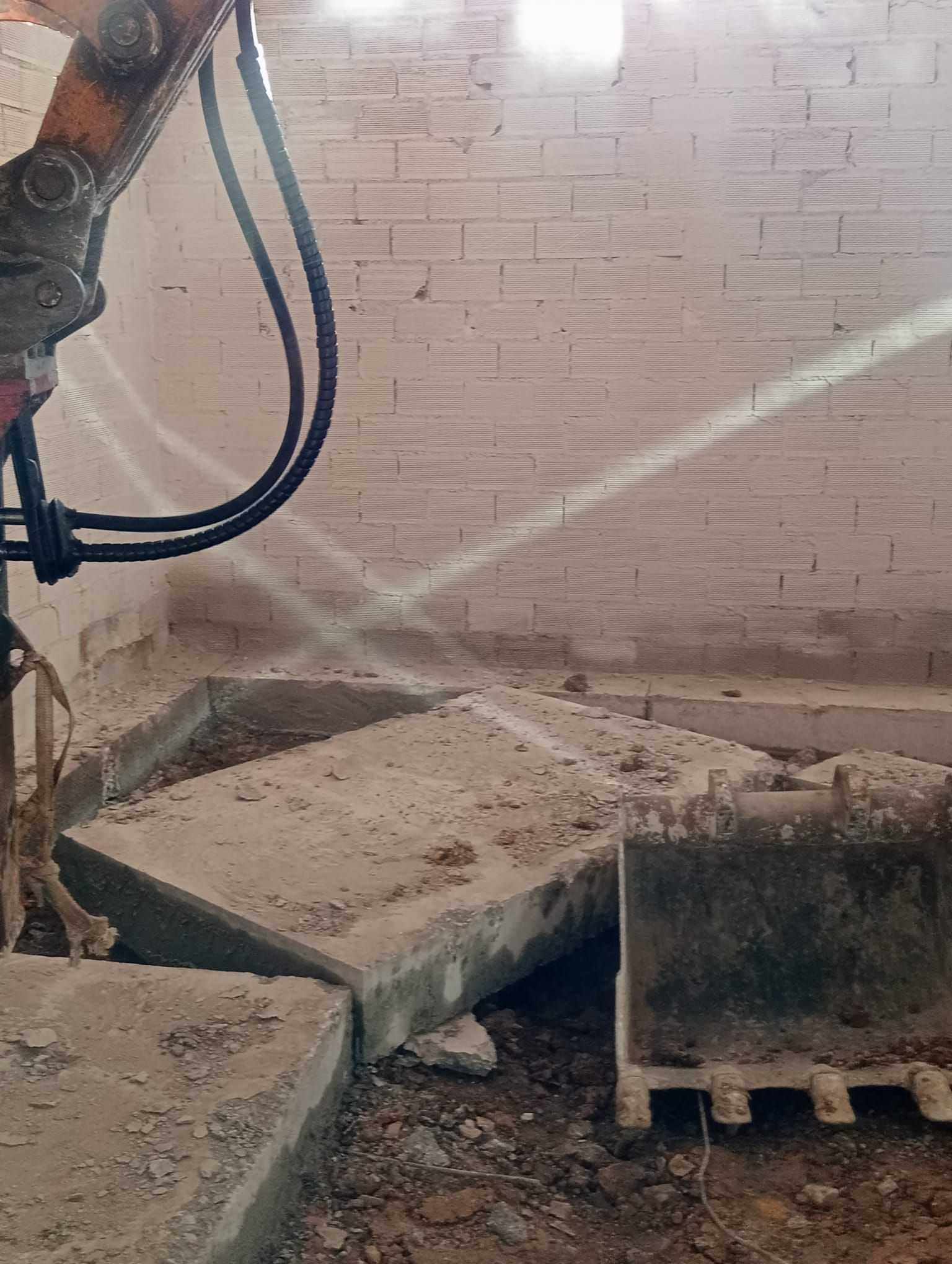 Obra en construcción polvorienta con losas de hormigón rotas y una manguera rociando agua contra una pared de ladrillos.