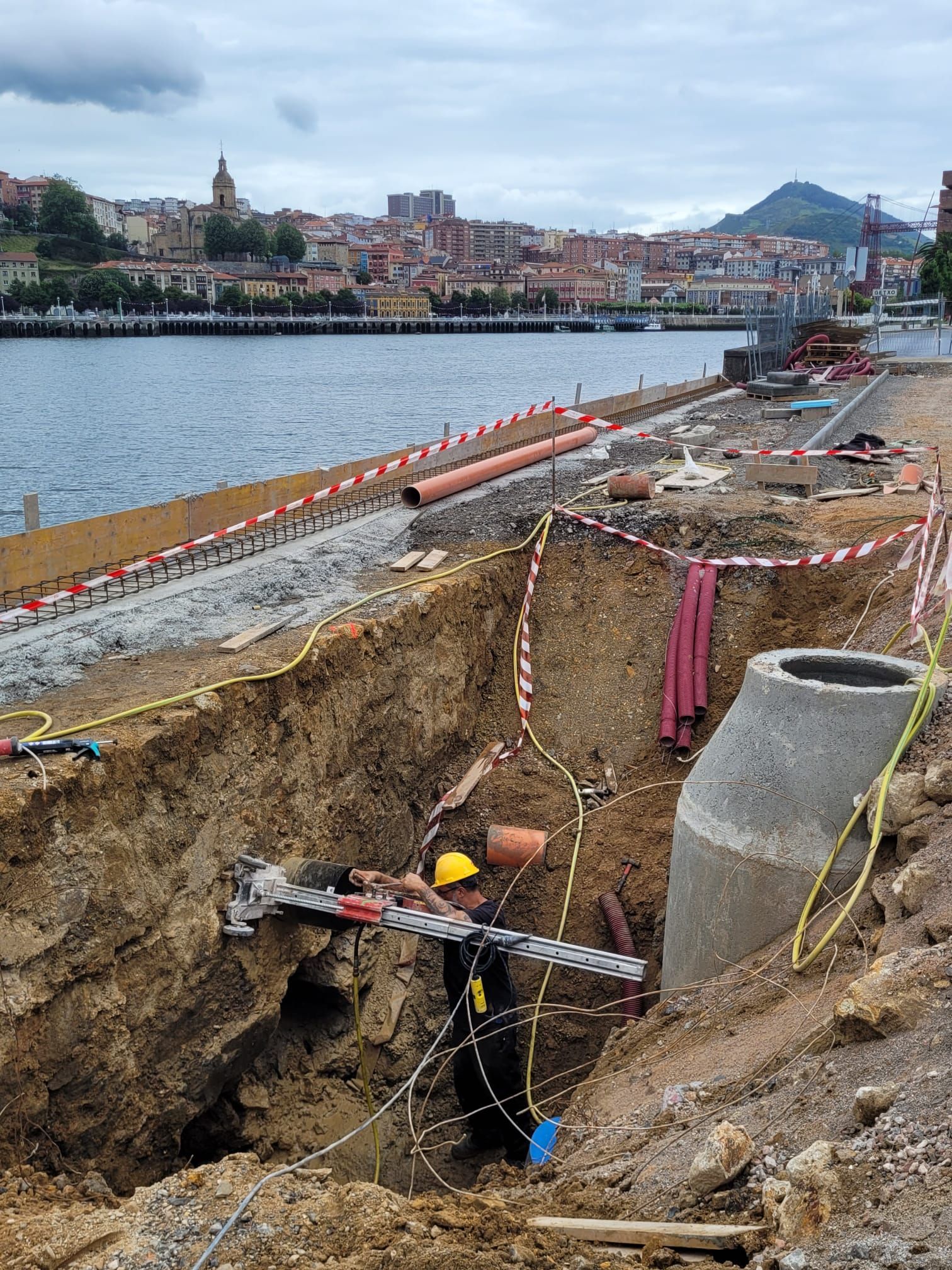 Un trabajador con casco entra en una zanja en una obra de construcción a orillas del río, con el perfil urbano de la ciudad al fondo.
