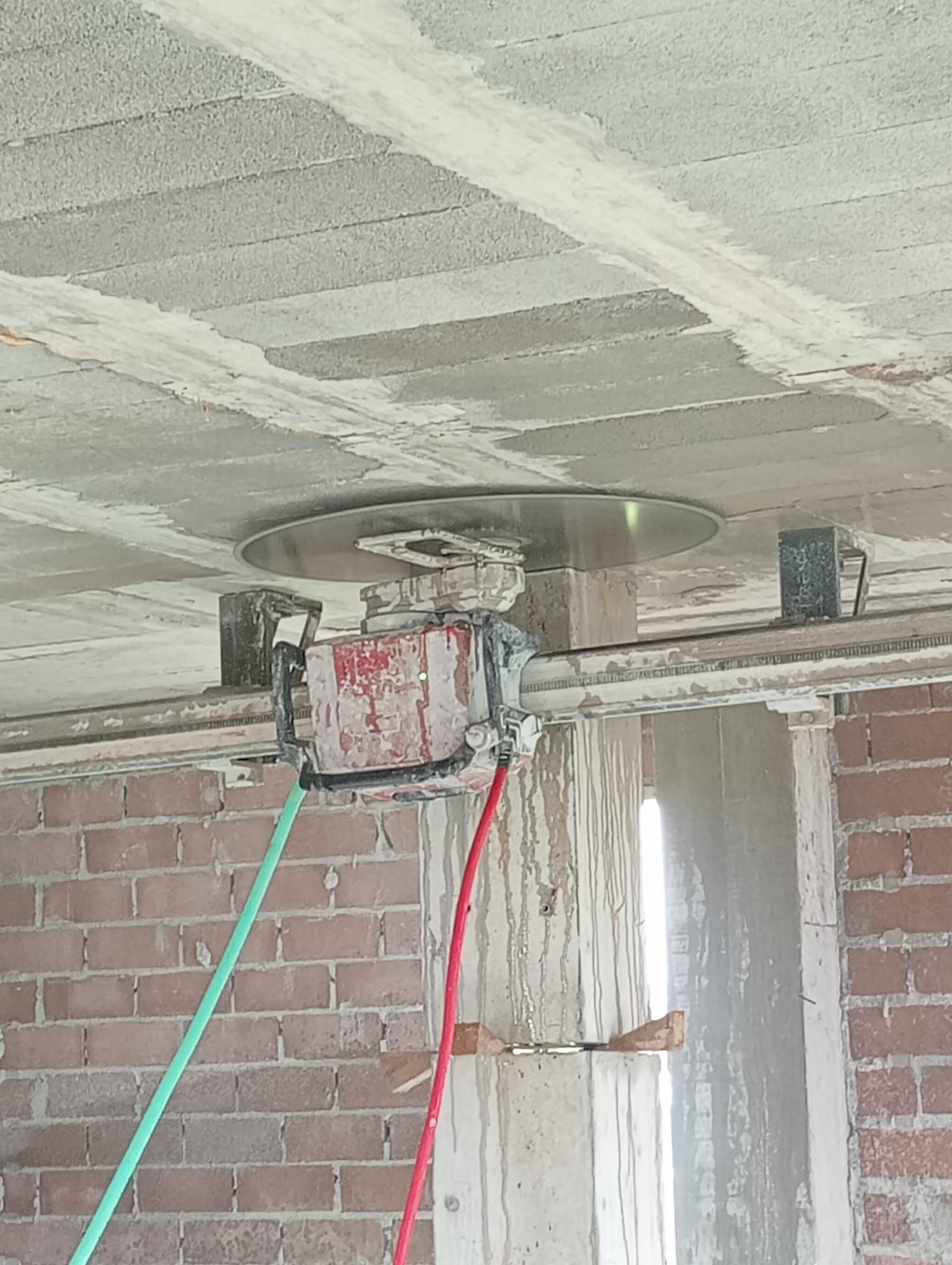 Techo de hormigón con una caja de conexiones circular, cables rojos y verdes colgantes y una pared de ladrillo en una habitación sin terminar.