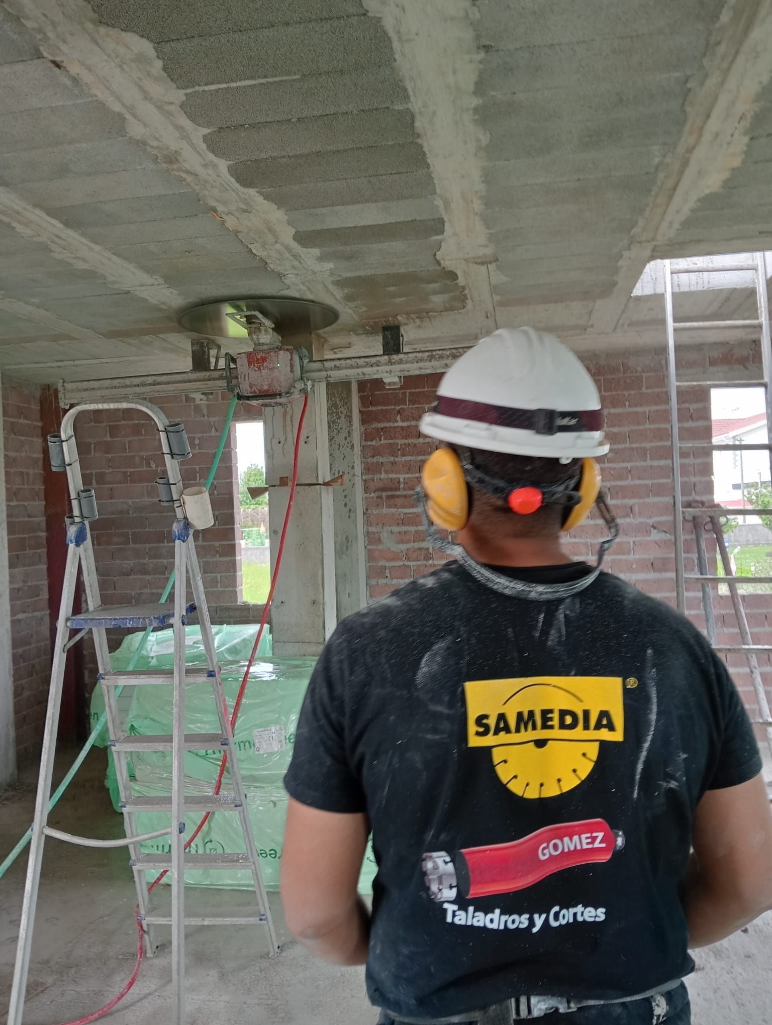Un trabajador con casco y protección auditiva se encuentra en el interior de una estructura de hormigón sin terminar, rodeado de escaleras y andamios.