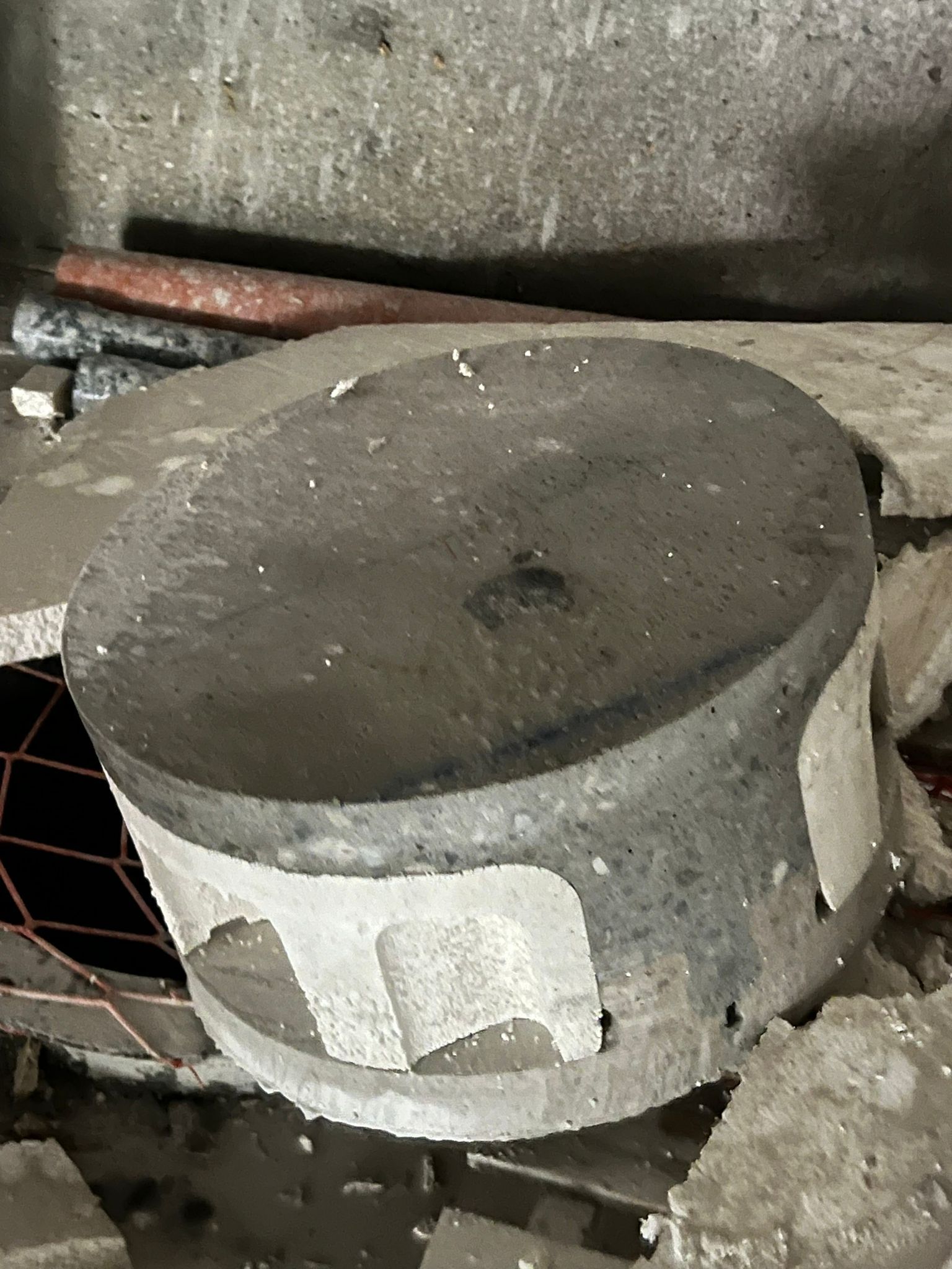 Cilindro de hormigón sobre una rejilla metálica, con la parte superior oscura y los laterales blancos desconchados, apoyado contra una pared rugosa.