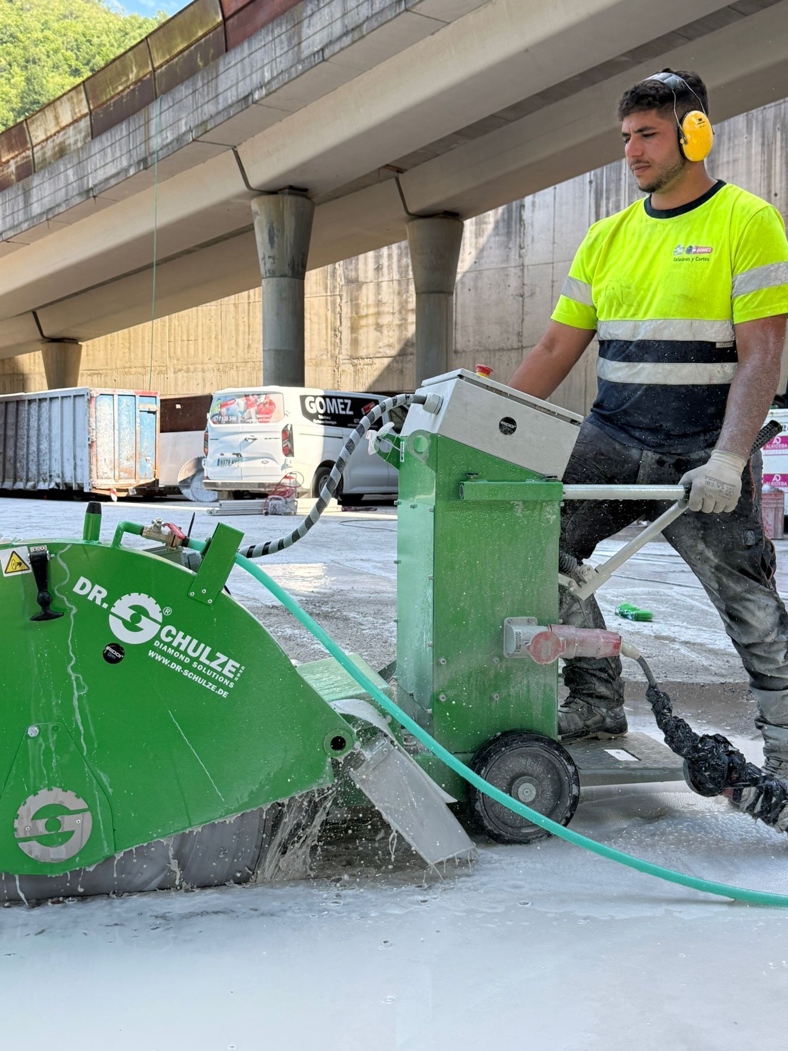 Un trabajador opera una sierra para pavimento verde con rociador de agua debajo de un paso elevado.