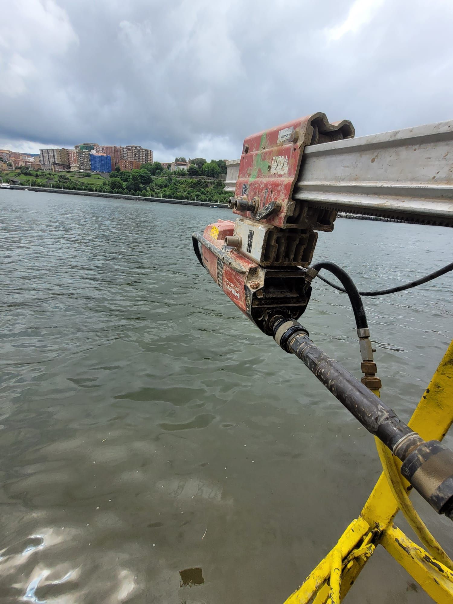 Maquinaria metálica desgastada montada sobre un puente que cruza un río, con edificios de la ciudad y un cielo nublado al fondo.