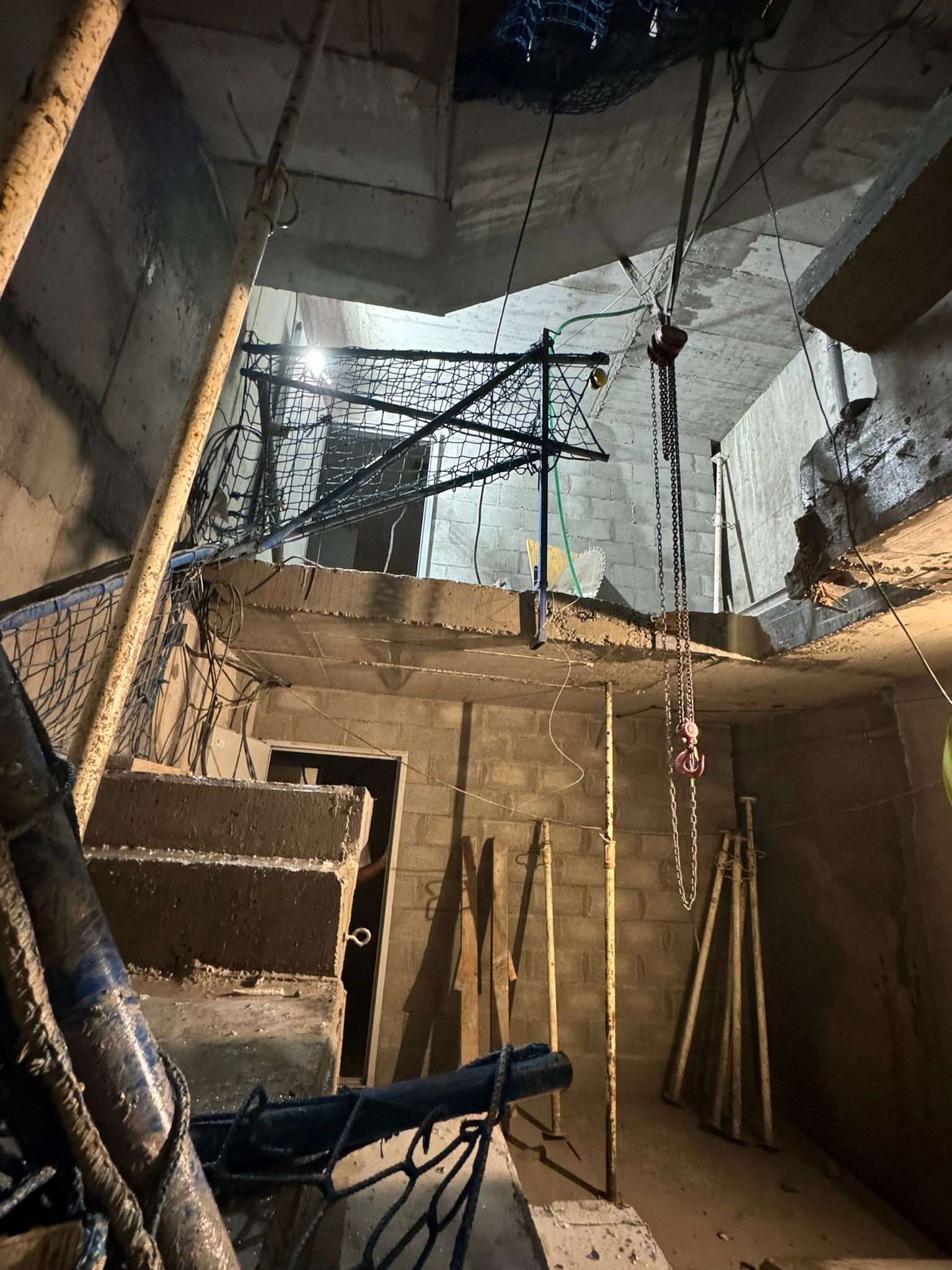 Una habitación de hormigón sin terminar, iluminada por el sol, con varillas de refuerzo a la vista, cables colgantes y una abertura para una escalera en el techo.