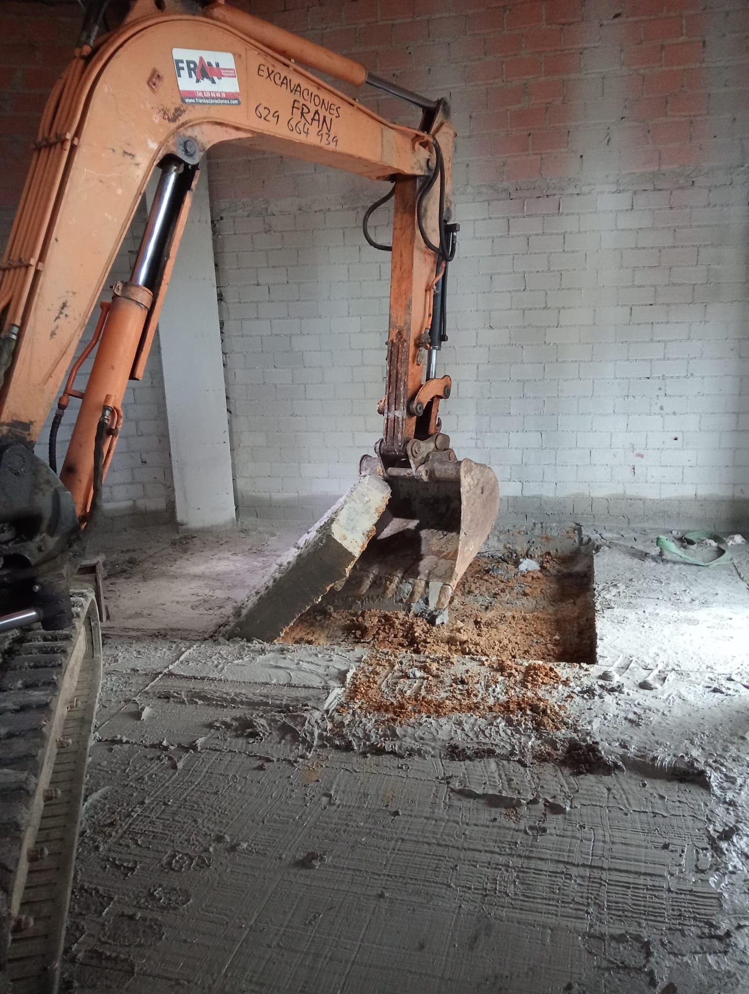 Una excavadora naranja demoliendo un piso de concreto dentro de una habitación con paredes de ladrillo.