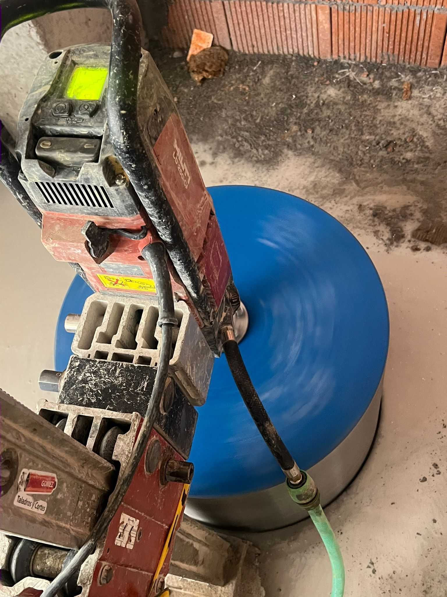 Un barril de plástico azul junto a una máquina pesada sobre un suelo de hormigón, con una manguera verde conectada.