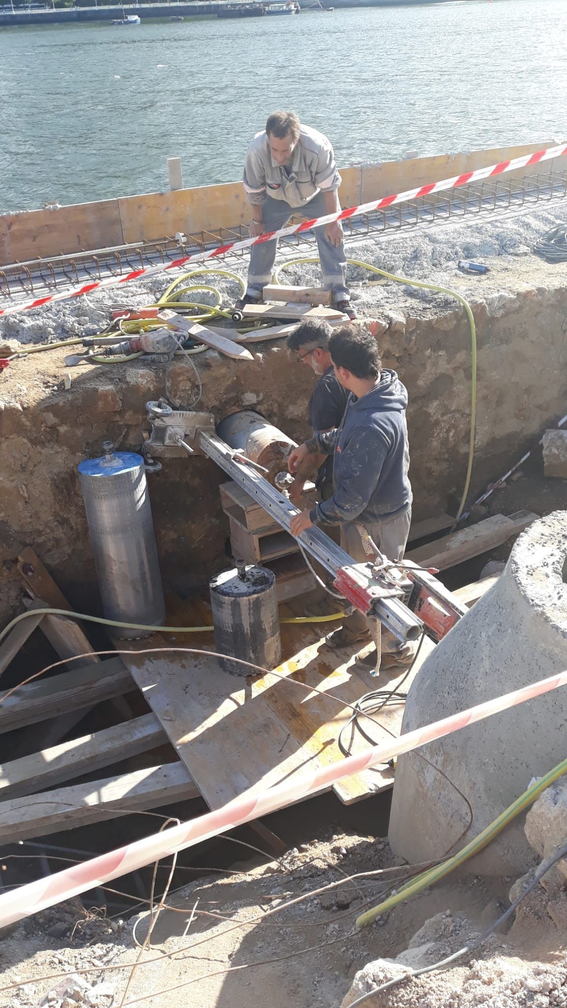 Obreros reparando un muro de contención de hormigón junto al agua, con herramientas y una escalera en el lugar.