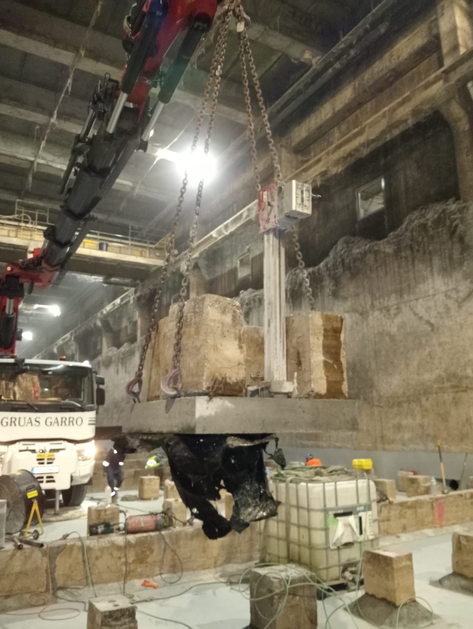 Obra subterránea con trabajadores, grúas, escombros y muros de hormigón en un túnel poco iluminado.
