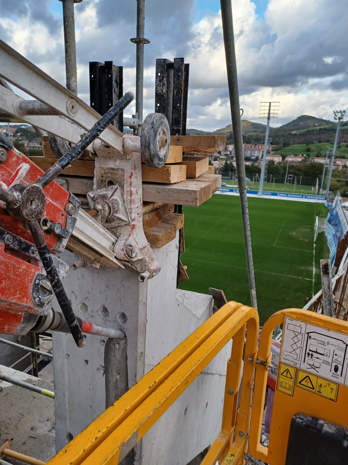 Plataforma de construcción con vistas a un campo de fútbol, con barandillas amarillas y herramientas en primer plano.