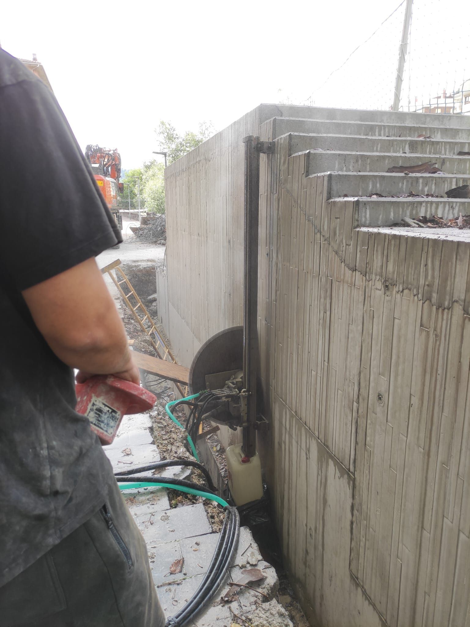 Una persona rocía agua contra una pared de hormigón en una obra de construcción al aire libre, en un entorno húmedo.