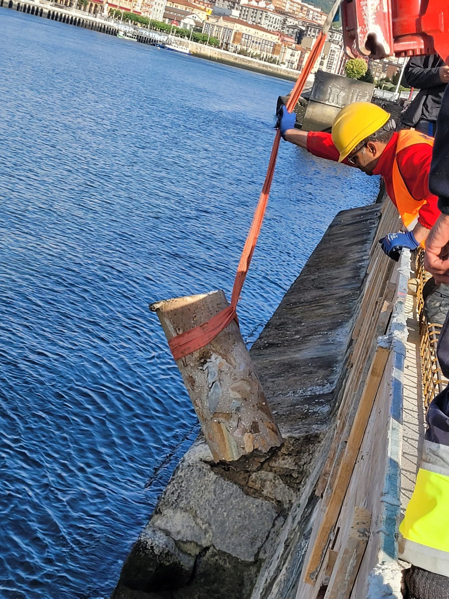 Trabajadores reparando un malecón, paleando escombros junto al agua azul cerca de un paseo marítimo.