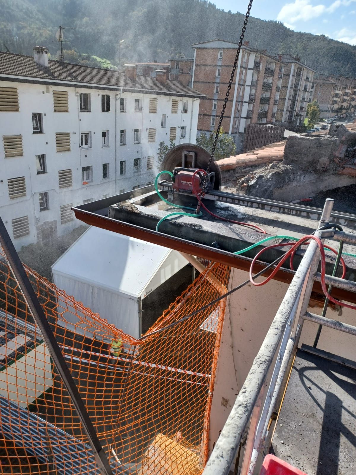 Obra en construcción en la azotea con un trabajador, red de seguridad naranja, cables y edificios de montaña al fondo.