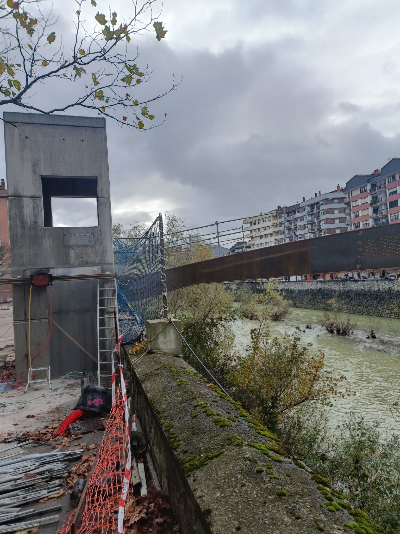 Paseo fluvial junto a una torre de hormigón, cielo nublado y un puente con edificios de apartamentos al fondo.