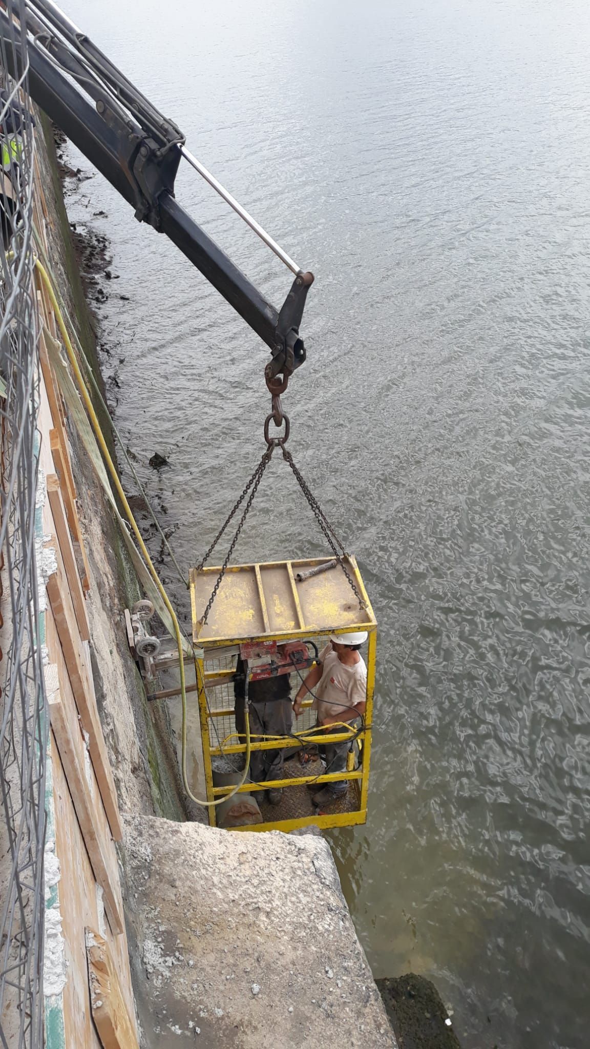 Un trabajador se encuentra en una cesta amarilla suspendida sobre el agua junto a un muro de hormigón, visto desde arriba.