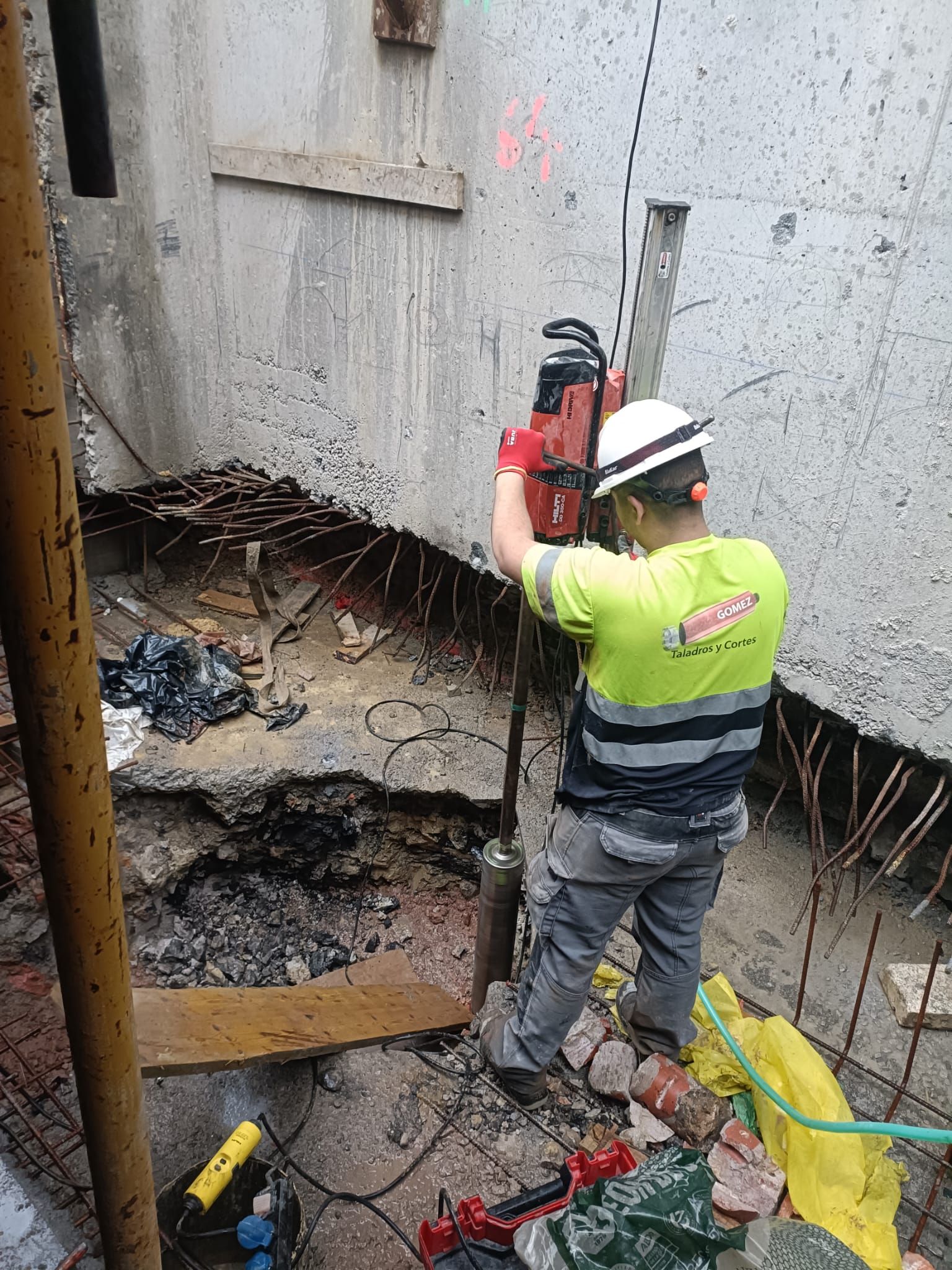 Un trabajador con equipo de seguridad fluorescente utiliza un martillo neumático en una zanja fangosa junto a un muro de hormigón.