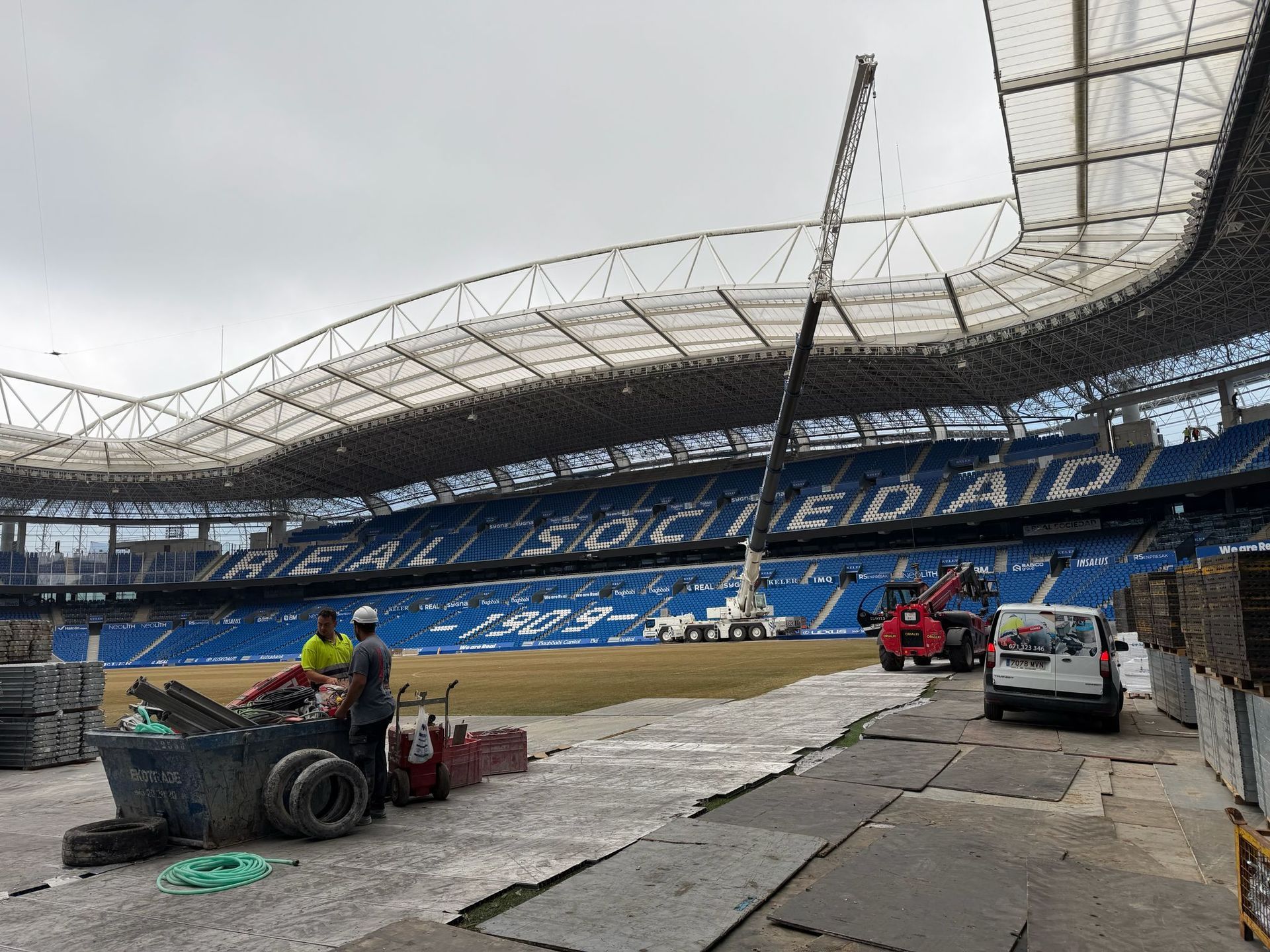 Trabajadores y maquinaria en el terreno de juego de un estadio bajo un techo parcialmente cubierto, con gradas al fondo.