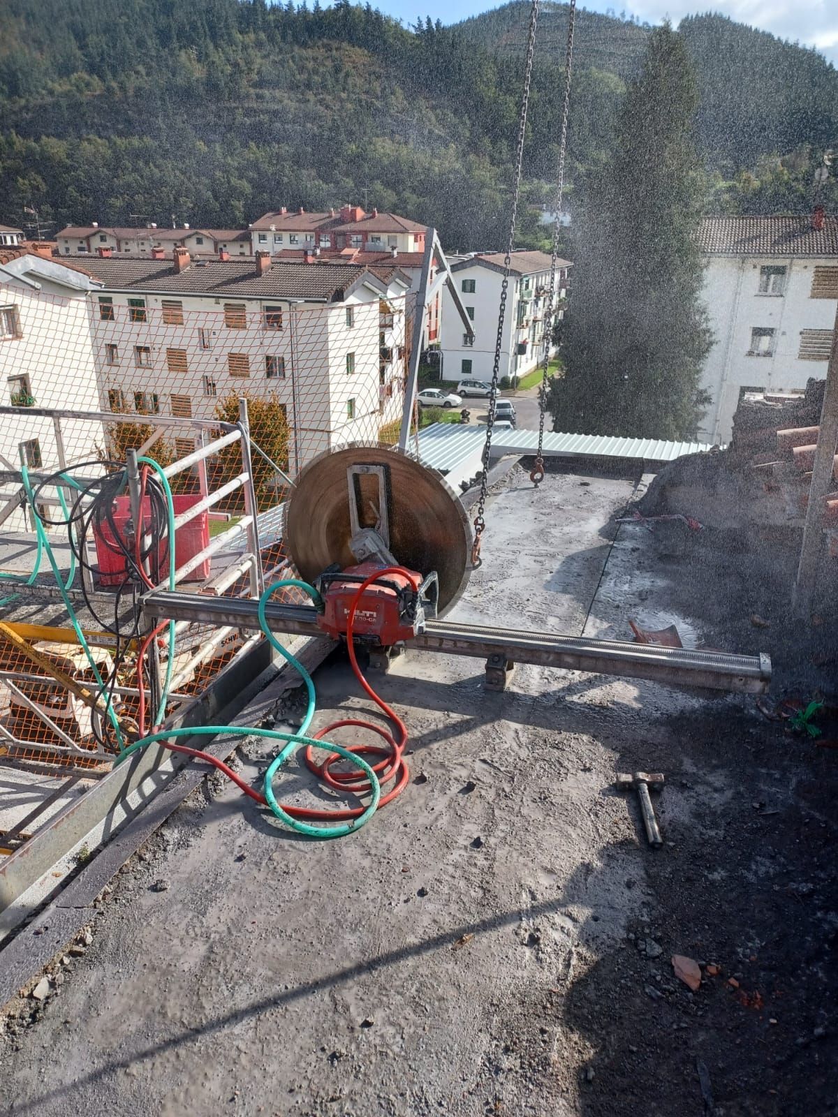Azotea en construcción con carrete de manguera, barandilla roja y pueblo de montaña al fondo.