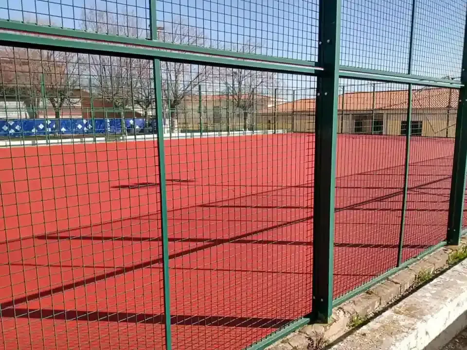 Cancha deportiva roja detrás de una valla metálica verde; cielo azul claro.