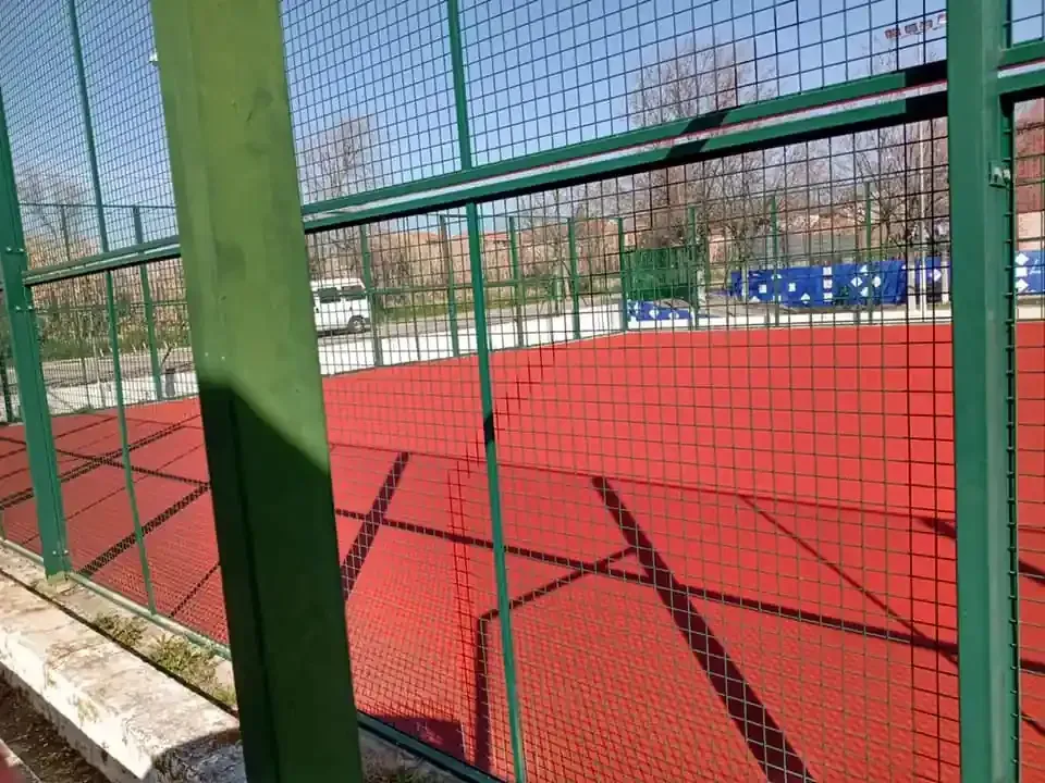 Pista de pádel roja tras una valla verde. Árboles y edificios al fondo. Luz solar brillante.