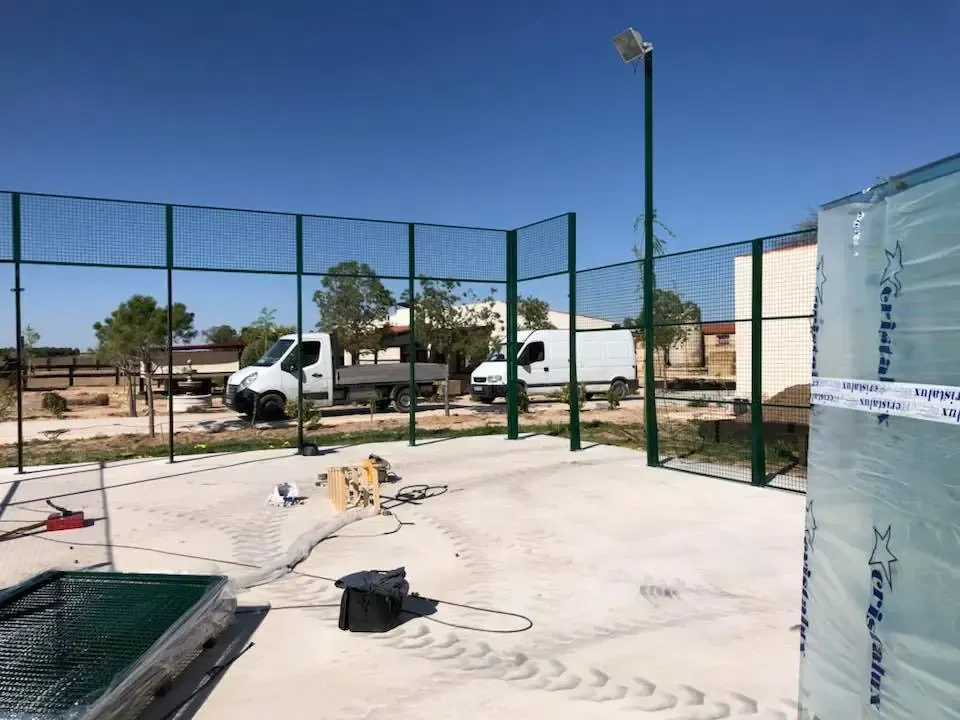 Pista de pádel en construcción, con vallado verde, un poste de luz y dos camiones en un día soleado.