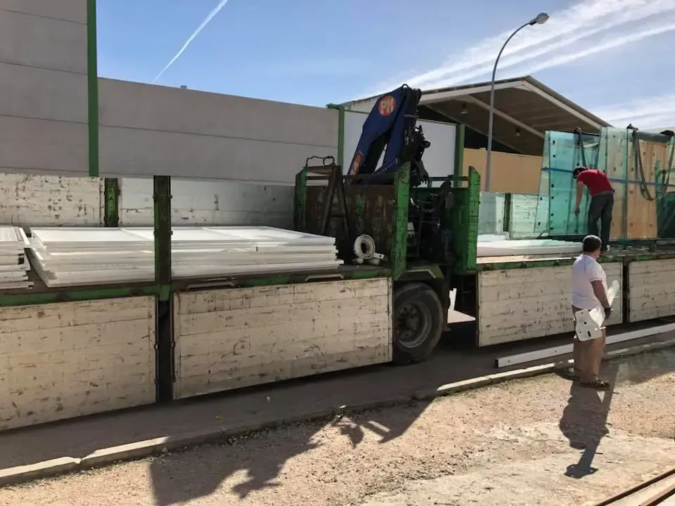 Camión con grúa cargando paneles de grandes dimensiones; dos personas trabajando al aire libre.