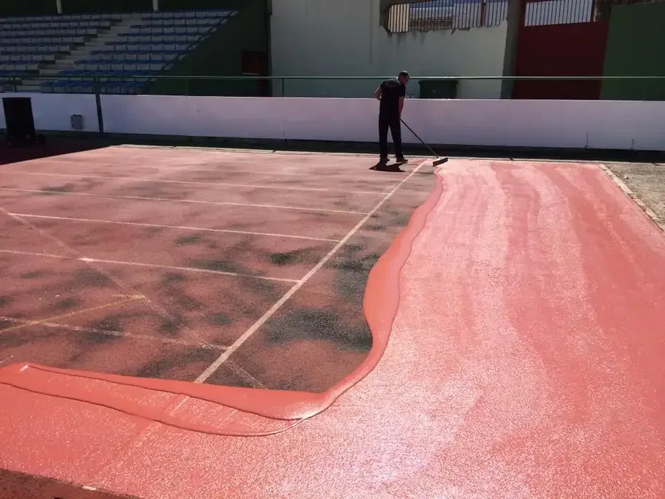 Persona aplicando una capa roja a una pista de atletismo con una herramienta de mango largo al aire libre.
