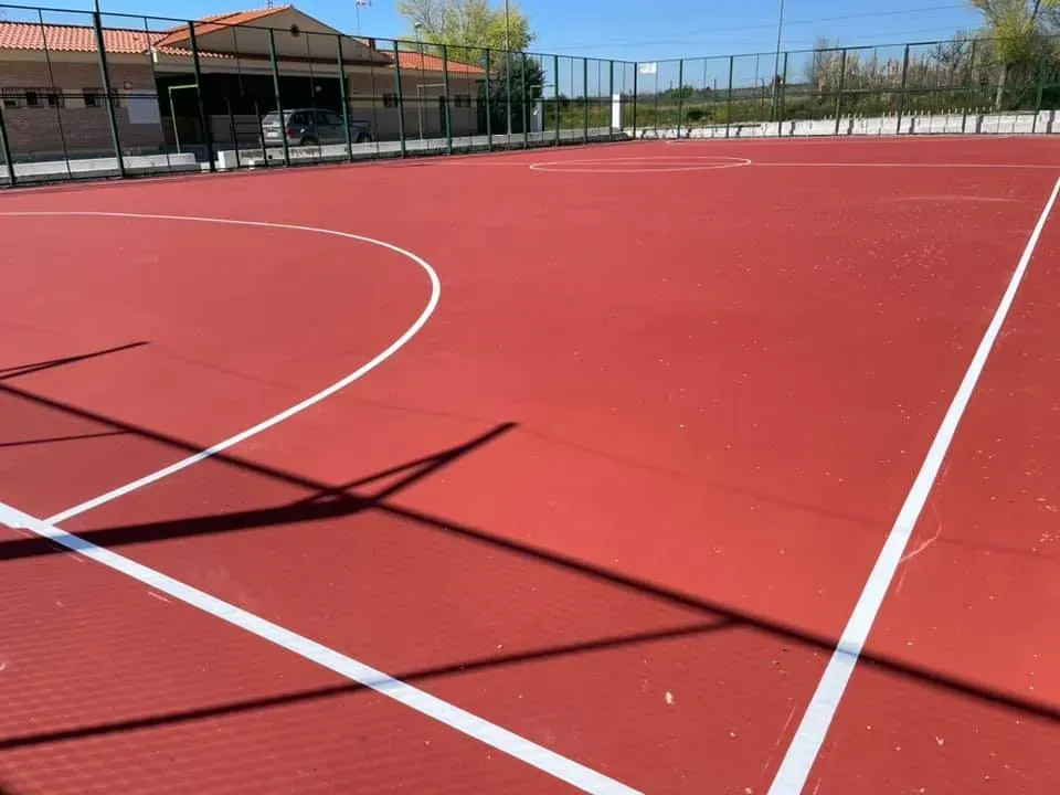 Cancha deportiva al aire libre de color rojo con líneas blancas, valla y edificio al fondo.
