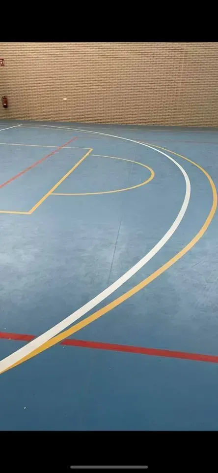 Cancha de baloncesto cubierta con suelo azul y líneas curvas blancas, amarillas y rojas. Pared beige al fondo.