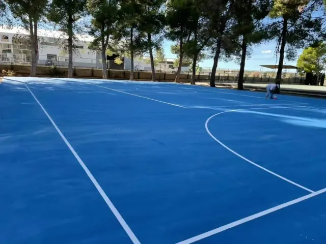 Cancha de baloncesto azul con líneas blancas, árboles al fondo, una persona pintando.