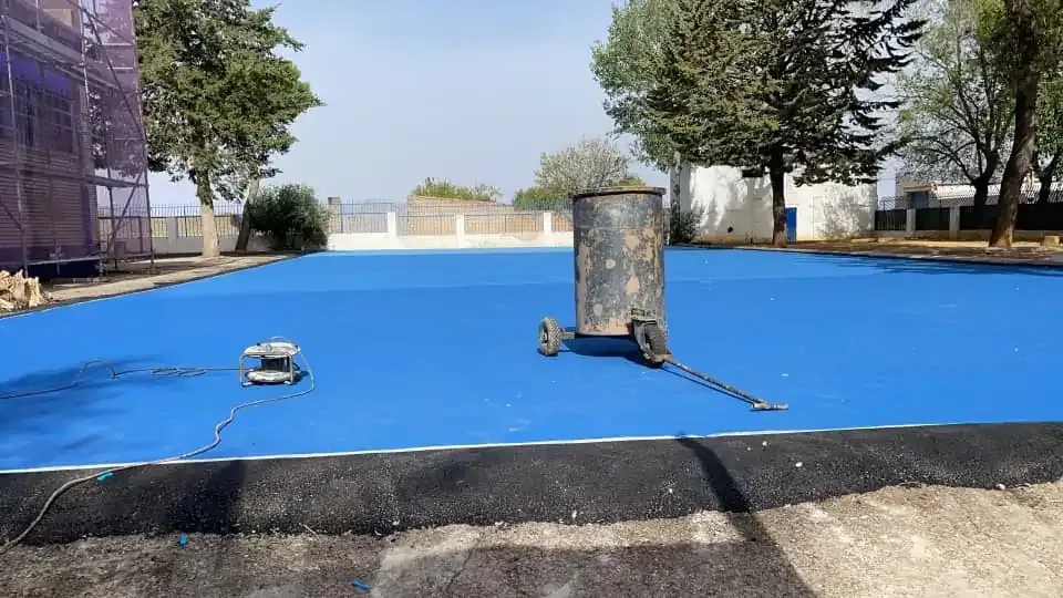 Área de superficie azul con un contenedor metálico y una herramienta encima. Árboles y un edificio al fondo.