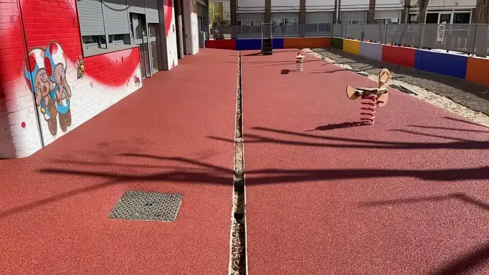 Superficie de patio de juegos roja con grietas, pared de graffiti y estructuras de juego.