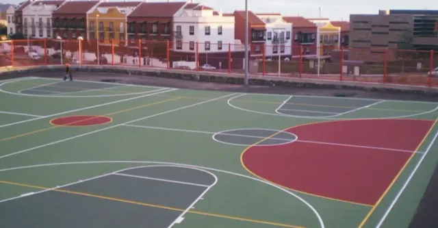 Cancha de baloncesto al aire libre con áreas pintadas de colores verde y rojo; casas al fondo.