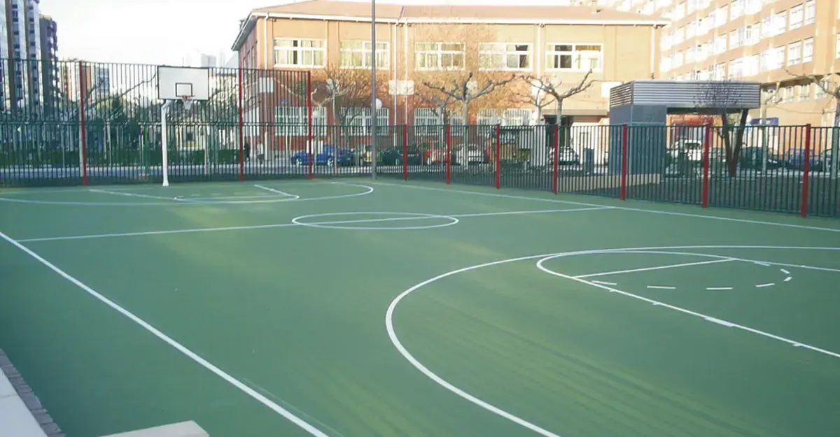 Cancha deportiva verde con lineas blancas y valla perimetral, junto a edificio.