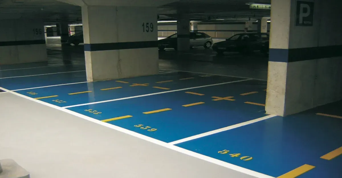 Estacionamiento con espacios pintados de azul, líneas blancas, números amarillos y pilares grises.