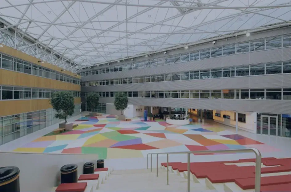 Gran atrio abierto con arte colorido en el piso, árboles, techo de vidrio y edificio de varios pisos.