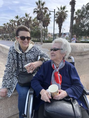 Dos personas en sillas de ruedas sonriendo al aire libre junto al mar, con palmeras al fondo.