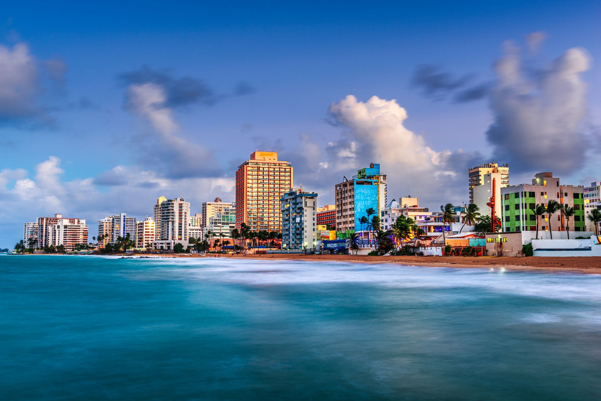City skyline with colorful buildings along a beach with turquoise water under a partly cloudy sky.