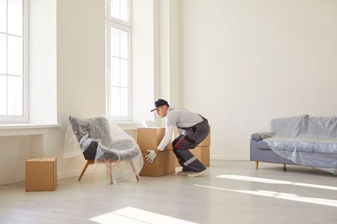 Umzugshelfer laden Kisten in einem hellen Raum mit abgedeckten Möbeln aus.